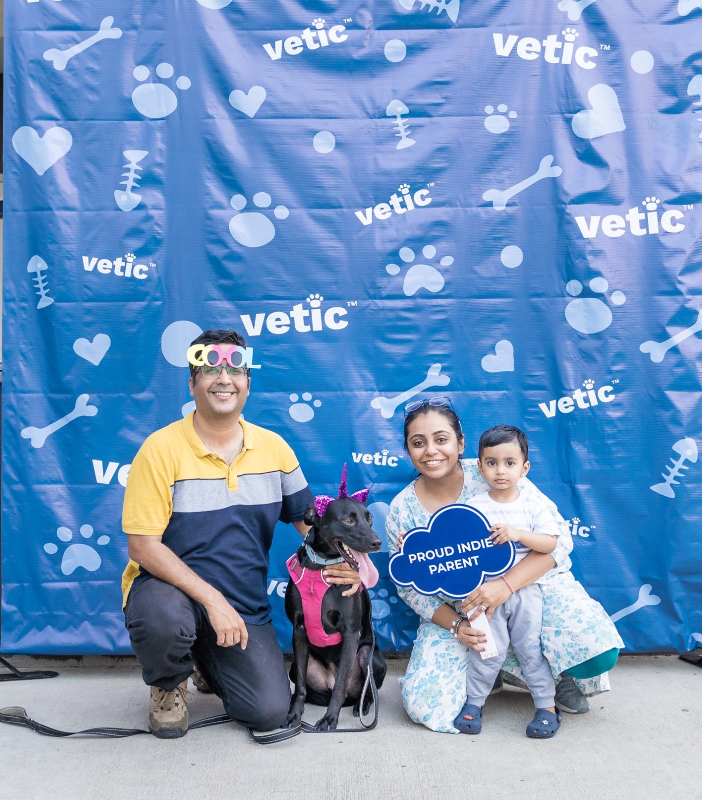 Photo of a family participating in an awareness camp. The family consists of the father, wearing photoshoot gear - a pair of COOL shades, the mum and their toddler holding a sign that says "proud indie parent" and at the center is their full black indie dog wearing a bright pink harness and matching unicorn headband.